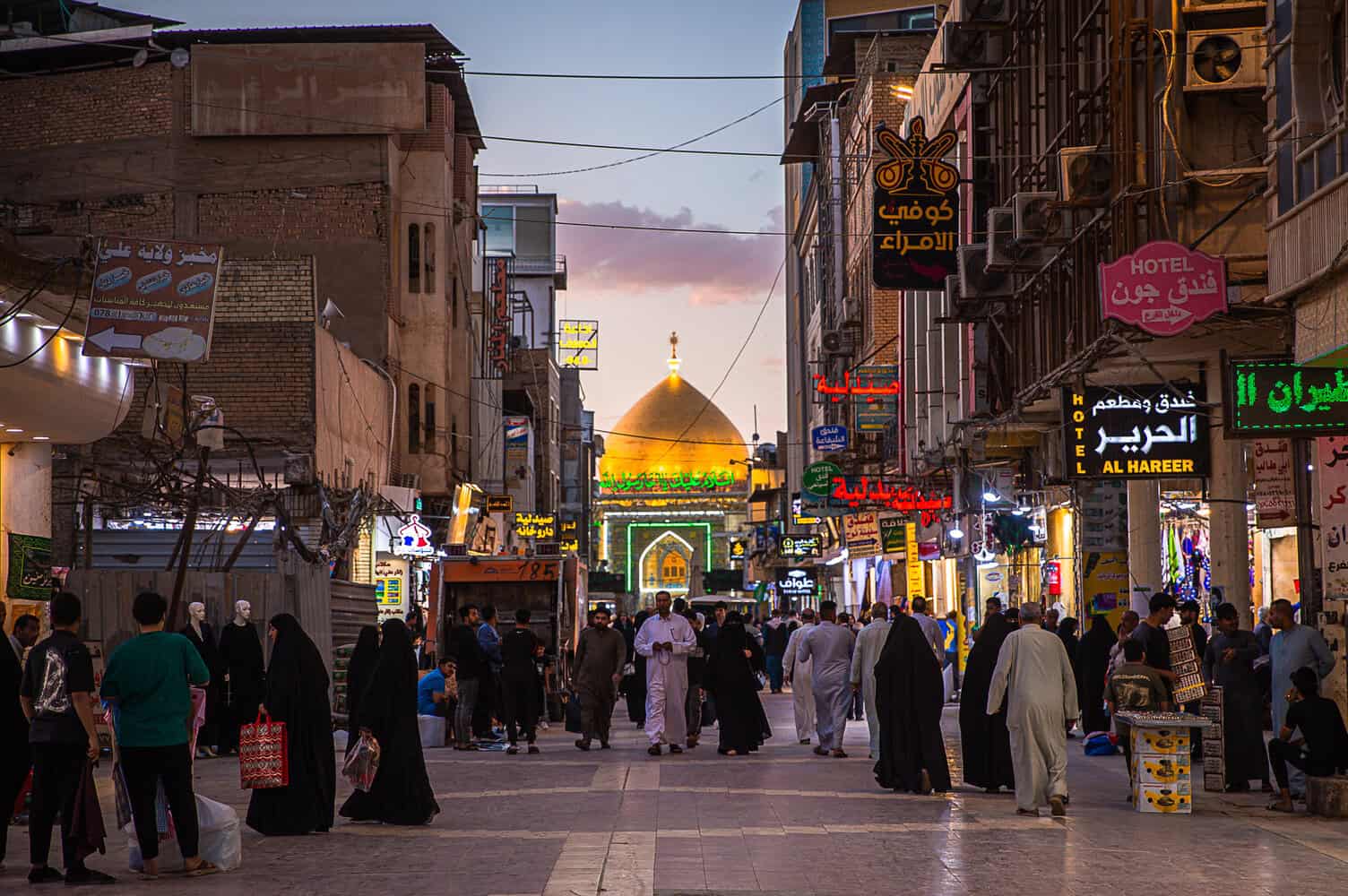Crowded pedestrian street leading to a glowing golden-domed shrine under a twilight sky.