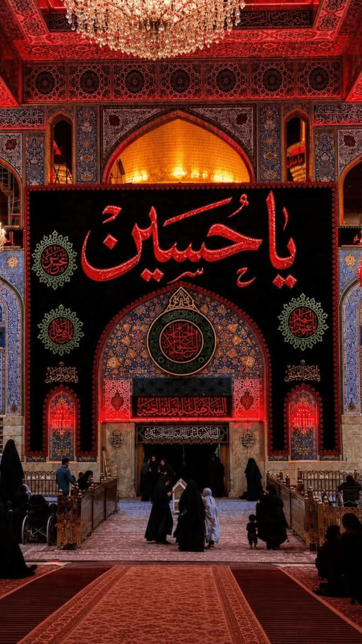 Ornate shrine interior with red lighting, a large 'Ya Hussain' banner, and people walking.