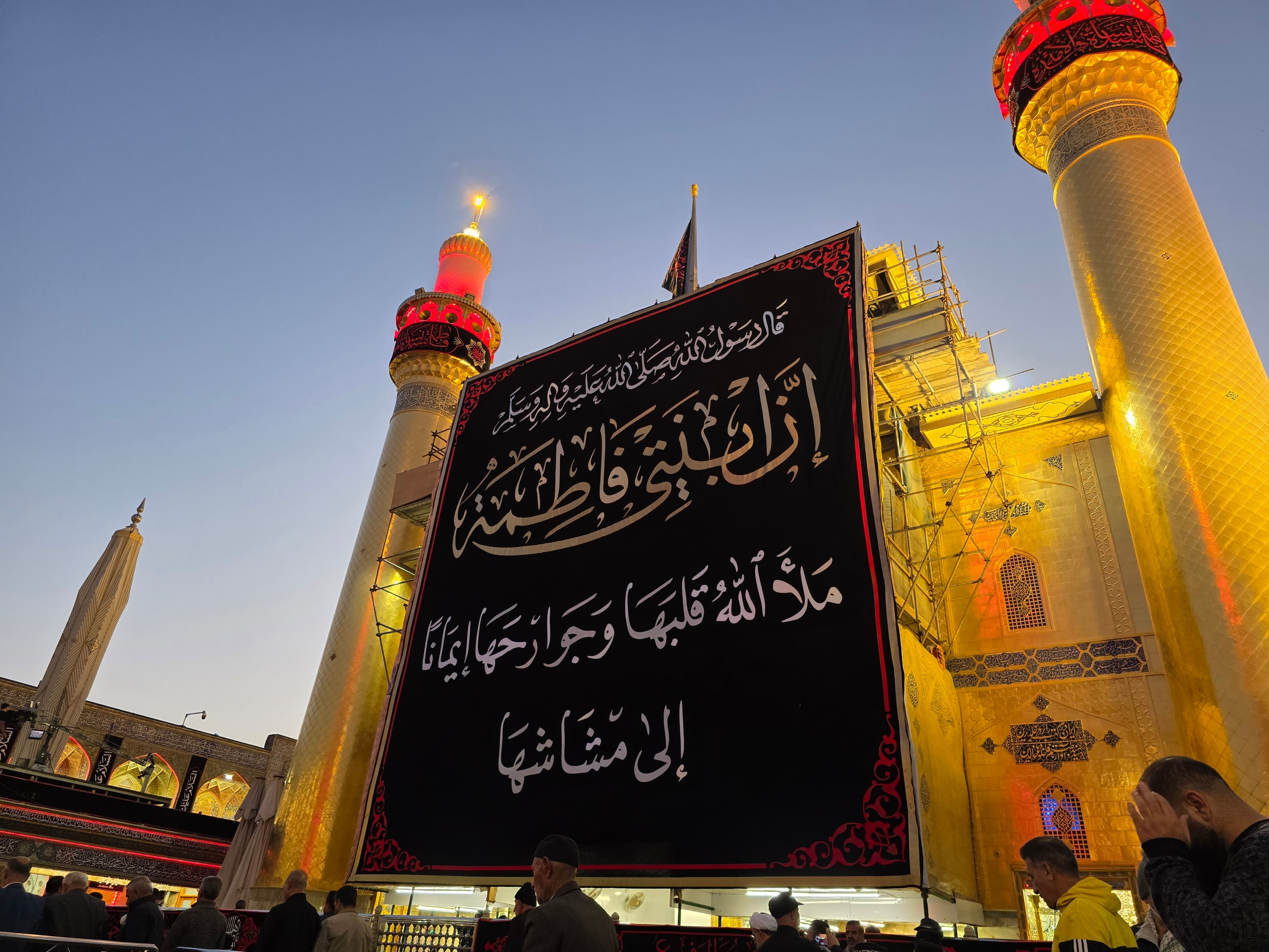 Large black banner with white Arabic calligraphy displayed before a golden mosque with illuminated minarets.