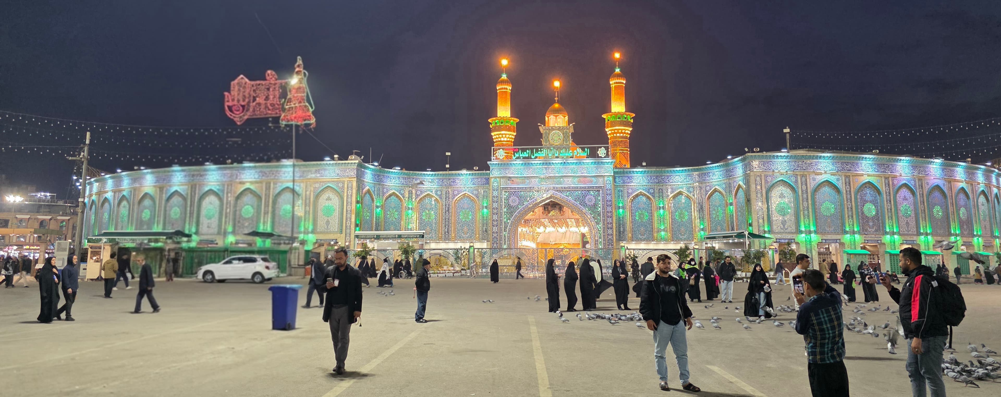 Grand shrine illuminated with green lights at night, featuring golden minarets and a crowded plaza.